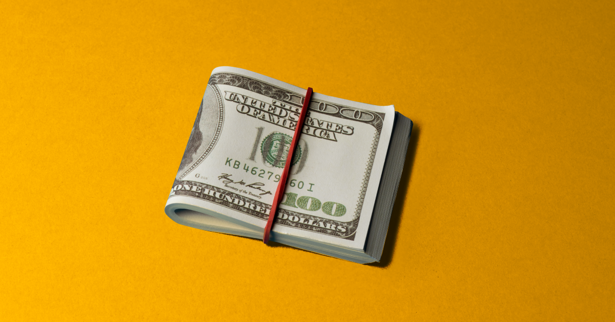 Folded up 100 dollar bill with a red rubber band on it on a yellow background. 