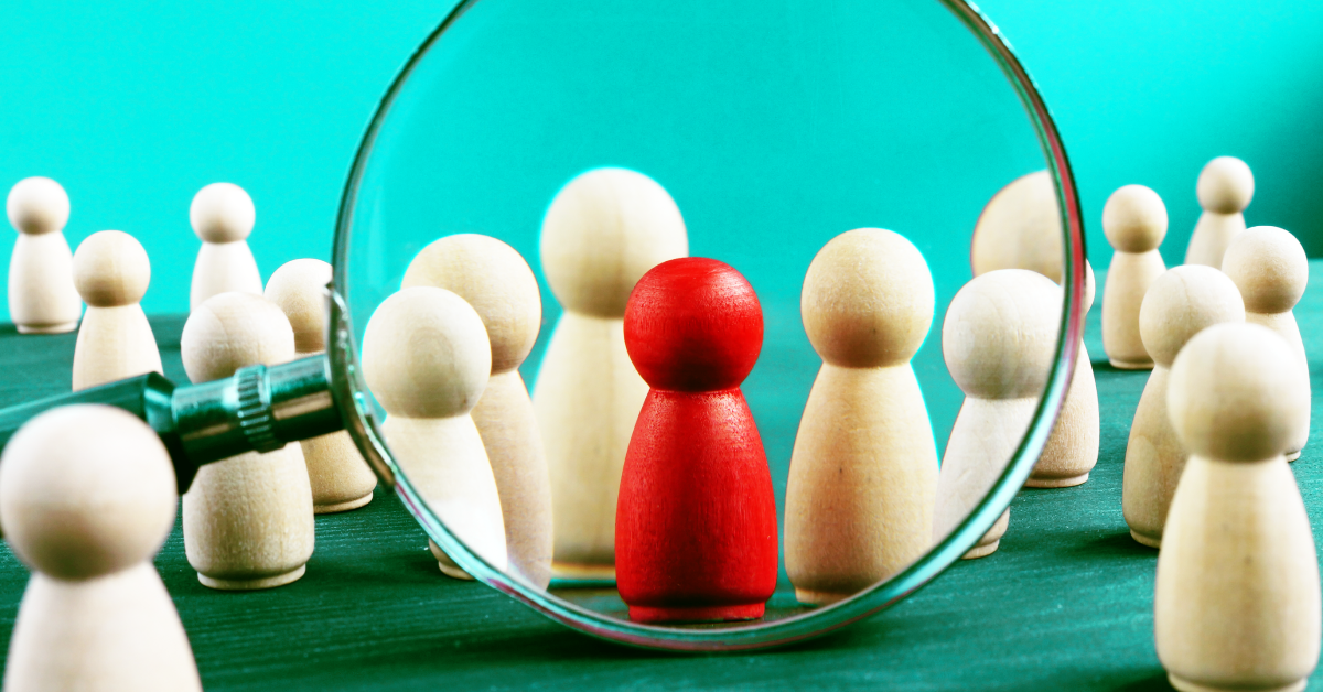 A magnifying glass looking at a collection of tan wooden figures and one red figure in the middle all on a sky blue background.